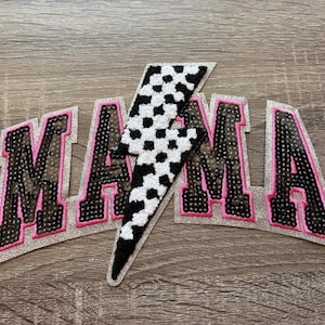 May include: Embroidered patches spelling "MAMA" with a black and white lightning bolt. The letters are black with pink trim and silver glitter backing. The lightning bolt is black and white checkered.