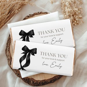 Op de afbeelding: Twee witte chocoladereepverpakkingen met zwarte strikillustraties en de tekst "THANK YOU for your love & support love, Emily". De repen worden gepresenteerd op een houten schijf.