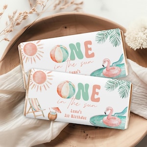May include: Two rectangular candy bar wrappers with a beach theme. The wrappers feature a sun, beach ball, and the text "ONE in the sun". A flamingo float and palm leaves are also shown. The text "Luna's 1st Birthday" is also included.