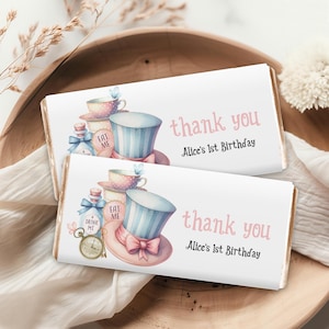 May include: Two white chocolate bar wrappers with an Alice in Wonderland theme. The wrappers feature illustrations of teacups, a bottle with the words "Drink Me", a hat with the words "Eat Me", and the text "thank you Alice's 1st Birthday".