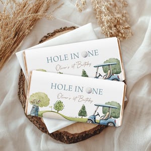 Pode incluir: Duas embalagens retangulares de barra de chocolate com um tema de golfe em aquarela. O texto diz HOLE IN ONE e Oliver's 1st Birthday. O design inclui um carrinho de golfe azul, uma bolsa com tacos, árvores verdes e uma bola de golfe.