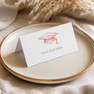 May include: A white folded card with a pink graduation cap illustration and the text "YOUR TEXT HERE". The card is on a beige plate with a gold rim, suggesting a graduation theme.