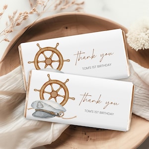 May include: Two white chocolate bar wrappers with a nautical theme. The top wrapper features a brown wooden ship's wheel and the text "Thank you Tom's 1st Birthday". The bottom wrapper features a brown wooden ship's wheel, a blue and white sailor's hat, and the text "Thank you Tom's 1st Birthday".