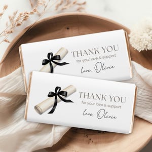 May include: Two white chocolate bar wrappers with the text "THANK YOU for your love & support love, Olivia." Each wrapper features a rolled diploma with a black ribbon. The bars are on a wooden tray.