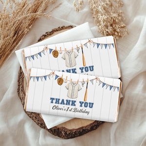 May include: Two rectangular chocolate bar wrappers featuring a white wood-plank background, blue pennant bunting, and baseball-themed illustrations including a glove, ball, jersey, whistle, and bat. The text reads THANK YOU Oliver's 1st Birthday.