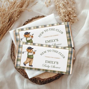 Pode incluir: Duas embalagens retangulares de barra de chocolate com uma borda xadrez verde, azul e bege. O centro exibe um urso de pelúcia vestido com roupa de golfe, com o texto "NEW TO THE CLUB - THANKS FOR COMING - EMILY'S Baby Shower" em verde escuro.
