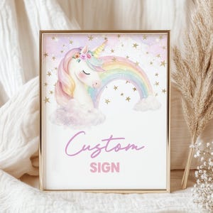 May include: A rectangular sign in a gold frame featuring a watercolor illustration of a white unicorn with a floral crown, a pastel rainbow, and gold stars against a soft purple and white background. The text Custom SIGN is printed in pink.