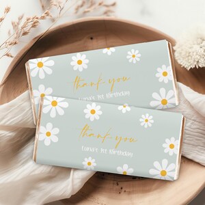 May include: Two light blue chocolate bar wrappers with white daisies and yellow centers. The wrappers have the text "thank you" in gold script and "Luna's 1st Birthday" in gold print.