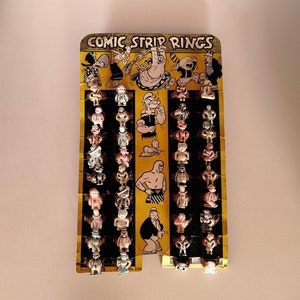 以下が含まれることがあります： 「コミックストリップリングス」という文字が印刷された黄色のカード。カードには、漫画のキャラクターの小さなカラフルなプラスチック製フィギュアのコレクションが掲載されています。