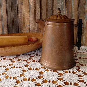 May include: A copper coffee pot with a black handle and a wooden lid finial sits on a white crocheted tablecloth. A wooden rolling pin rests in a wooden bowl. The background is a wooden wall.