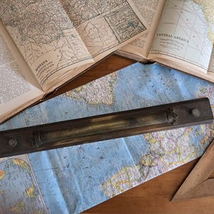 May include: A vintage wooden and brass parallel ruler with a sliding mechanism. The ruler is lying on top of a map of the world, with a wooden triangle ruler in the bottom right corner.