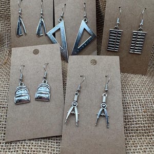 May include: A collection of silver-toned earrings displayed on brown cardstock. The earrings feature various designs, including a stapler, a triangle ruler, an abacus, a backpack, and a compass. The earrings are hanging on silver hooks.