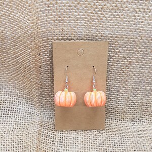 May include: A pair of pumpkin-shaped earrings with silver hooks. The pumpkins are orange with green stems and white stripes.