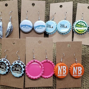 May include: Assortment of 10 pairs of earrings made from repurposed bottle caps. The bottle caps feature various logos and designs, including Seagram's, Coca-Cola, Frosty Rootbeer, Sun Shine Soda, and NB. The earrings are silver and have a hook closure.