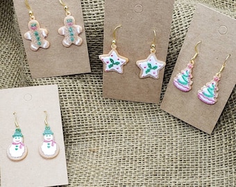 May include: Four pairs of Christmas-themed earrings displayed on brown card stock. The earrings are gingerbread men, stars with holly, and Christmas trees.