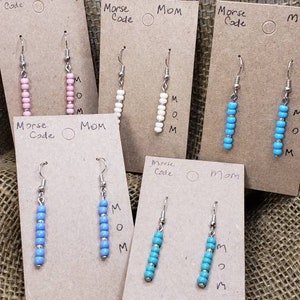 Könnte beinhalten: Fünf Paar Ohrhänger auf braunen Pappkarten mit der Aufschrift Morse Code MOM. Jedes Paar besteht aus einem vertikalen Strang kleiner Perlen in Rosa, Weiß, Hellblau, Mittelblau oder Türkis, die an silberfarbenen Ohrhaken befestigt sind.