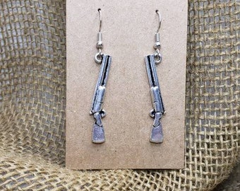 May include: Silver-colored shotgun earrings with a detailed design, hanging from silver-tone ear wires. The earrings are displayed on a brown card against a textured burlap background.