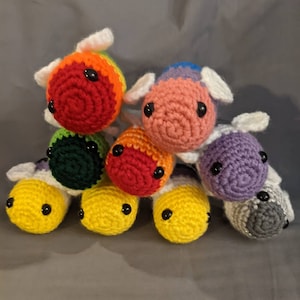 May include: A collection of handmade crochet cow toys in various colors, including yellow, green, orange, red, purple, and rainbow. Each cow has black button eyes and white ears. The toys are stacked together, showcasing their vibrant hues.