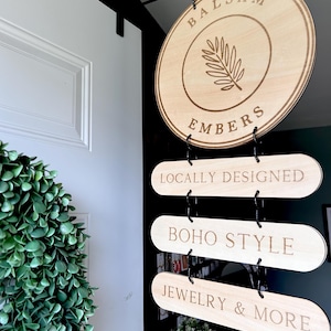 Pode incluir: Uma placa de madeira com o texto "Balsam Embers" e um desenho de folha. Abaixo da placa, h&aacute; tr&ecirc;s placas menores que dizem "Locally Designed", "Boho Style" e "Jewelry & More".