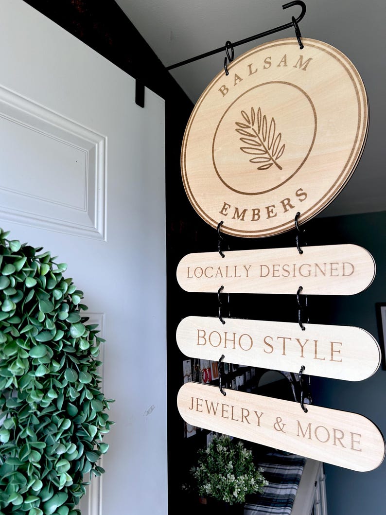 Pode incluir: Uma placa de madeira com o texto "Balsam Embers" e um desenho de folha. Abaixo da placa, h&aacute; tr&ecirc;s placas menores que dizem "Locally Designed", "Boho Style" e "Jewelry & More".