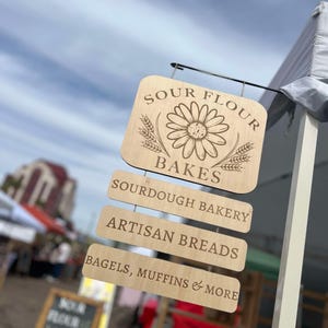 Pode incluir: Uma placa de madeira com o texto "Sour Flour Bakes" e um gr&aacute;fico de margarida. A placa tamb&eacute;m tem tr&ecirc;s placas menores penduradas abaixo, que dizem "Sourdough Bakery", "Artisan Breads" e "Bagels, Muffins & More".