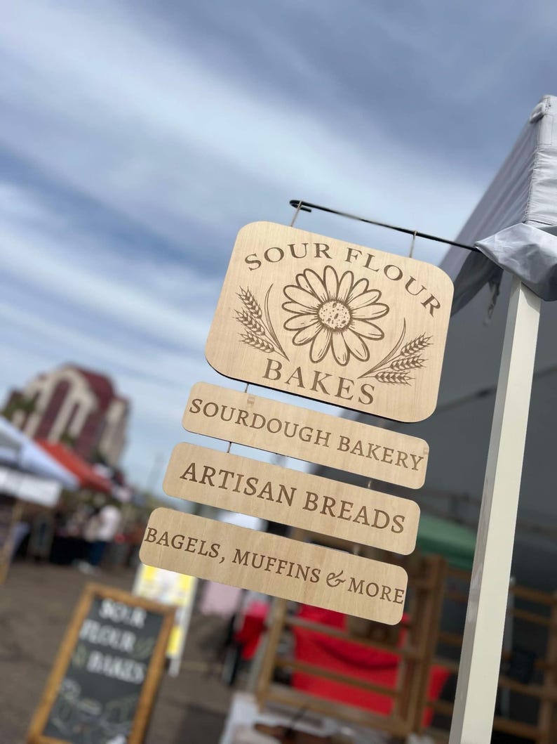 Pode incluir: Uma placa de madeira com o texto "Sour Flour Bakes" e um gr&aacute;fico de margarida. A placa tamb&eacute;m tem tr&ecirc;s placas menores penduradas abaixo, que dizem "Sourdough Bakery", "Artisan Breads" e "Bagels, Muffins & More".