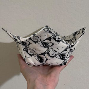 May include: A white fabric bowl with a black and white print of stormtrooper faces. The bowl is folded and held in a hand.