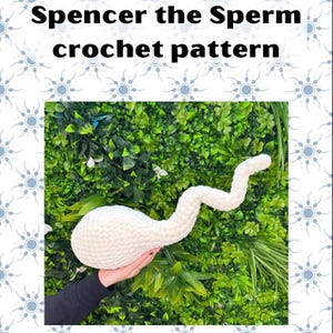 May include: A white, crocheted sperm-shaped plush toy against a backdrop of green foliage. The text "Spencer the Sperm crochet pattern" is displayed at the top of the image. The plush toy is held by a person.