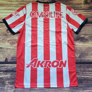 May include: Red and white striped soccer jersey with the word "OMNILIFE" printed on the back. The jersey also has the logos for "AKRON" and "CNP" printed on the back.