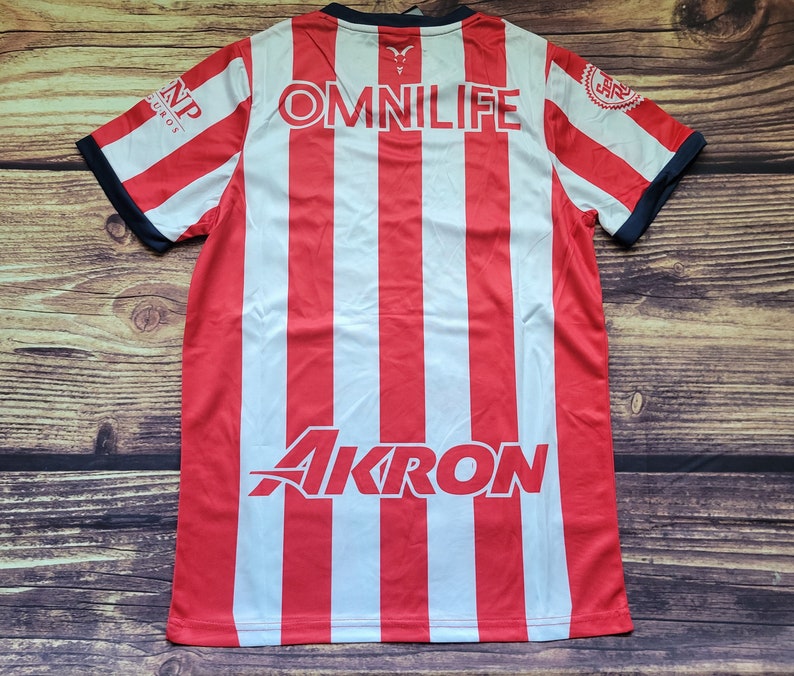 May include: Red and white striped soccer jersey with the word "OMNILIFE" printed on the back. The jersey also has the logos for "AKRON" and "CNP" printed on the back.