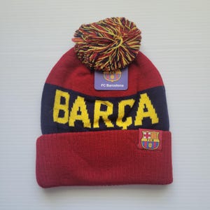 May include: Red and blue knit beanie with a yellow pom pom and a yellow "BARCA" logo. The beanie also has a small FC Barcelona logo patch.