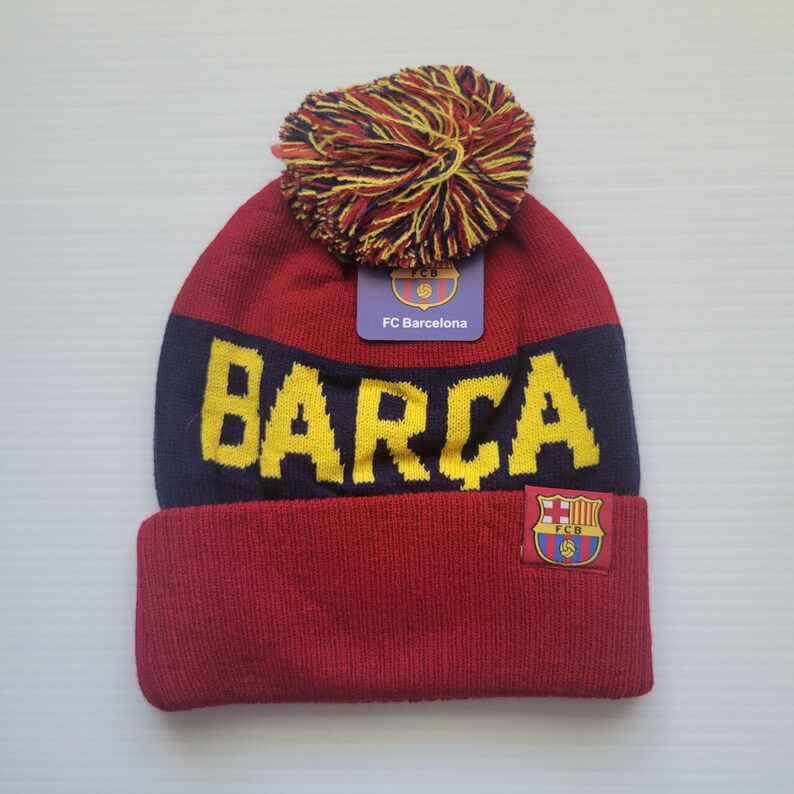May include: Red and blue knit beanie with a yellow pom pom and a yellow "BARCA" logo. The beanie also has a small FC Barcelona logo patch.