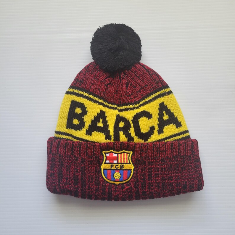 May include: A red and yellow knit beanie with a black pom pom on top. The beanie has the word "BARCA" in black letters on the front and a red and blue FC Barcelona logo.