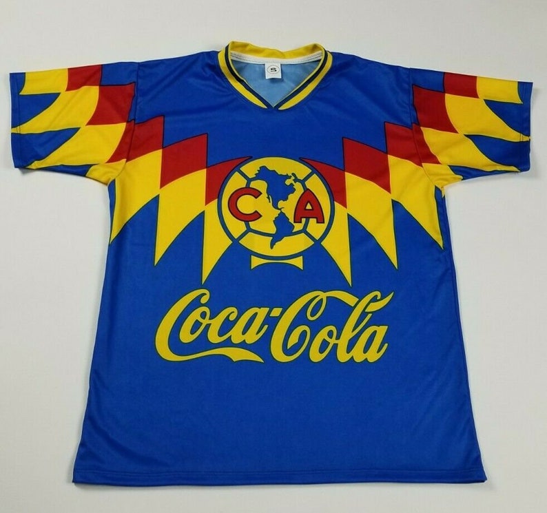 May include: Blue soccer jersey with yellow and red accents. The jersey features a large logo with the letters "CA" and a soccer ball. The text "Coca-Cola" is printed in yellow on the front of the jersey.