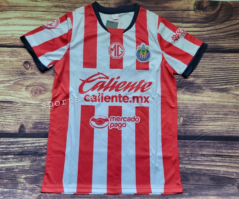 May include: Red and white striped soccer jersey with the team logo and sponsor logos. The jersey has the text "Caliente caliente.mx" on the front.