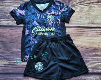 May include: A black and blue soccer jersey with a pink and blue floral pattern. The jersey has a V-neck and short sleeves. The jersey has the text "Caliente caliente.mx" on the front. The jersey is paired with black soccer shorts. The shorts have a green and yellow logo on the left leg.
