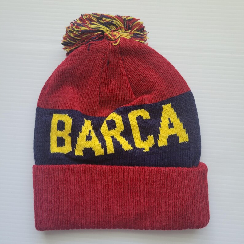 May include: Red and blue knit beanie with a yellow pom pom and the word "BARCA" in yellow letters.