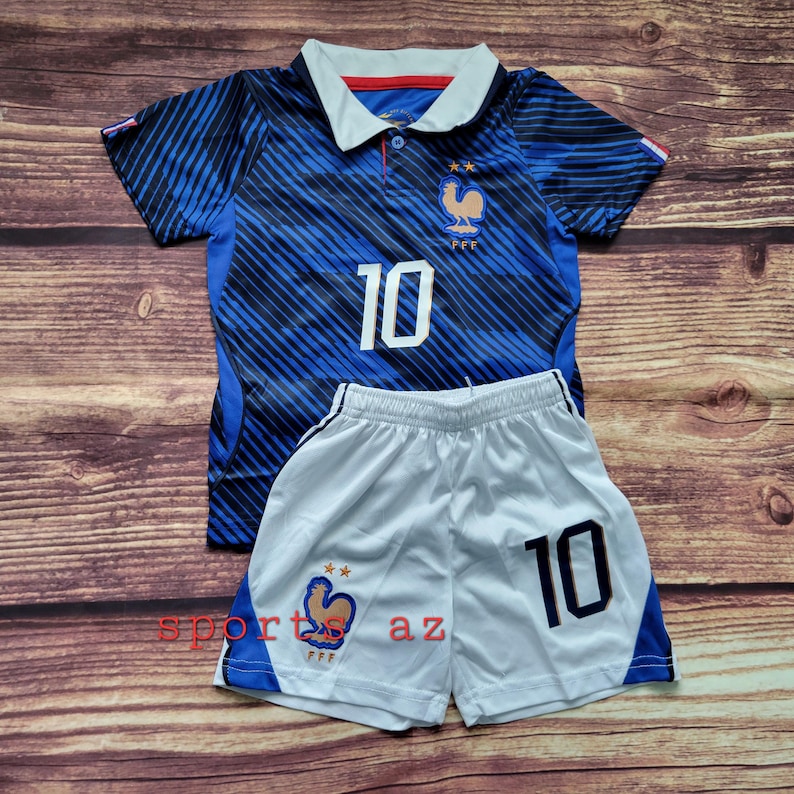 May include: A blue and black striped soccer jersey with a white collar, the number 10, and a gold rooster emblem with two stars. Paired with white soccer shorts featuring blue side panels, the number 10, and a matching gold rooster emblem.