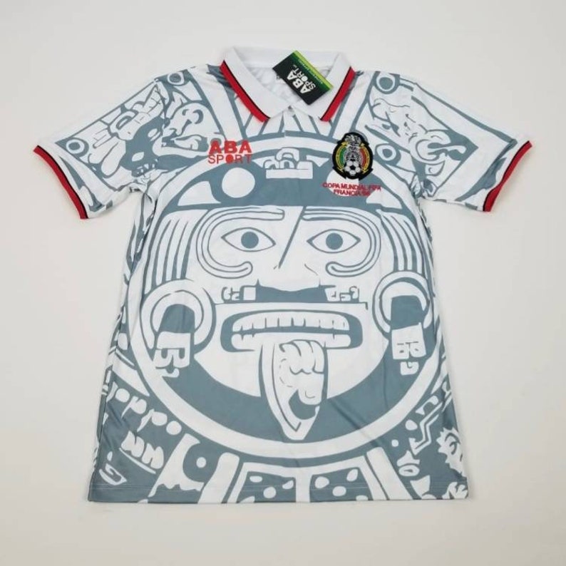 Op de afbeelding: Wit en grijs voetbalshirt met een groot Azteeks ge&iuml;nspireerd ontwerp op de voorkant. Het shirt heeft een witte kraag met rode bies, korte mouwen met rode manchetten en het "ABA SPORT" logo. Een label is aan de kraag bevestigd.