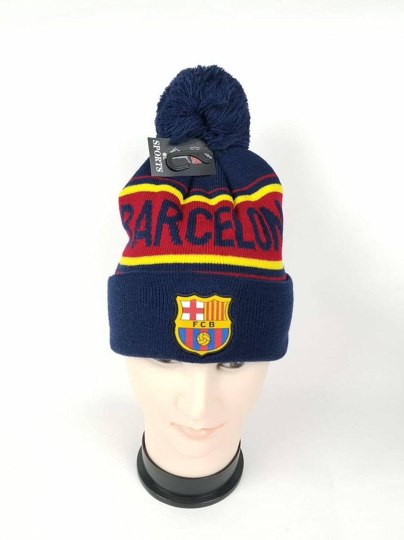 May include: A blue knitted beanie with a pom pom on top. The beanie has a red and yellow stripe pattern and a FC Barcelona logo with the text "FCB".