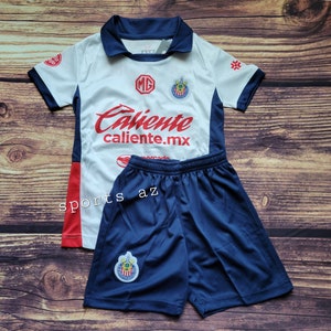 May include: White and blue soccer jersey with red lettering that says "Caliente caliente.mx". The jersey has a red and white stripe on the side and a team logo on the chest. The jersey is paired with blue soccer shorts that have the team logo on the left leg.