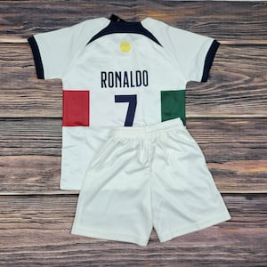May include: White soccer jersey with a blue and black collar, a yellow circle with a soccer ball graphic, and the text "RONALDO" and the number "7" on the back. The jersey has red and green accents on the sides. The jersey is paired with white soccer shorts.