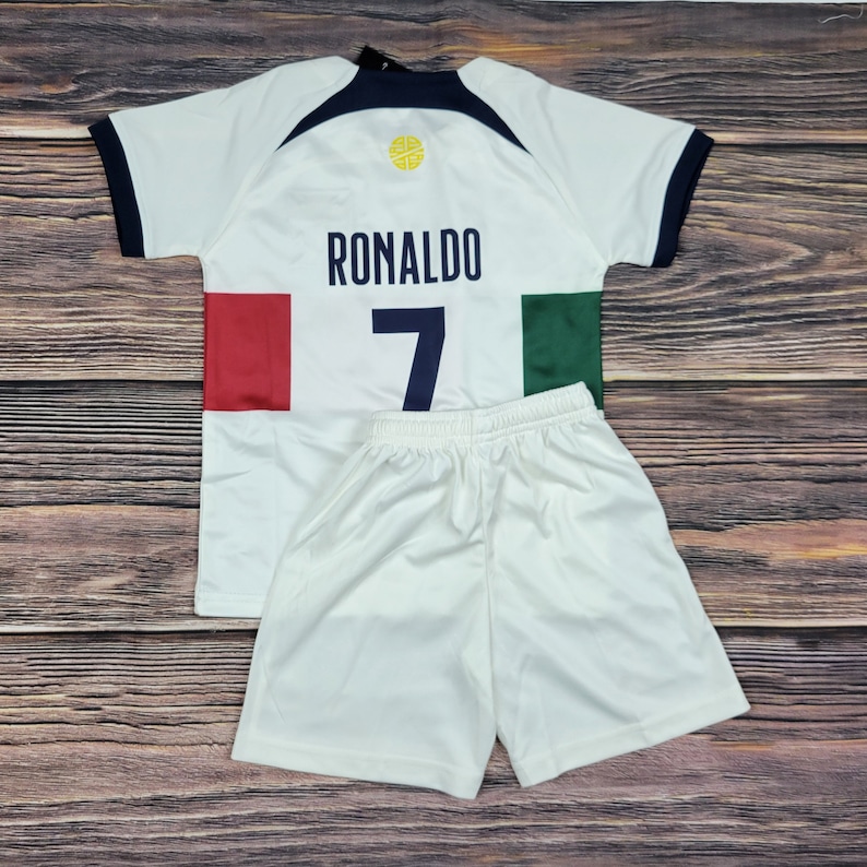 May include: White soccer jersey with a blue and black collar, a yellow circle with a soccer ball graphic, and the text "RONALDO" and the number "7" on the back. The jersey has red and green accents on the sides. The jersey is paired with white soccer shorts.