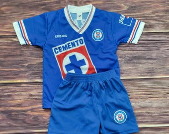 May include: A blue soccer jersey and shorts set. The jersey has a V-neck with white and red trim, and the word "CEMENTO" is printed on the front. The shorts are solid blue, and the set has the number "2" on the jersey.