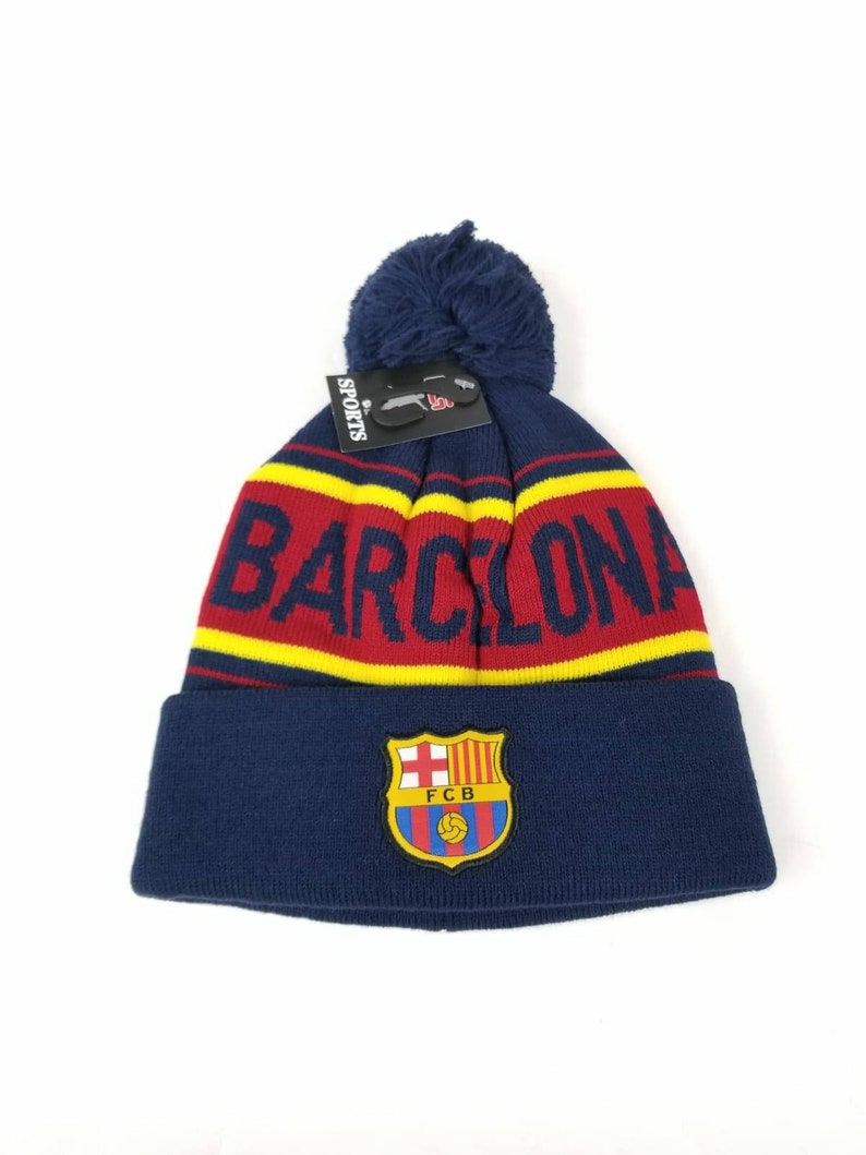May include: A blue knit beanie with a red and yellow stripe and the word "BARCELONA" in white. The beanie has a pom-pom on top and a FC Barcelona logo on the front.