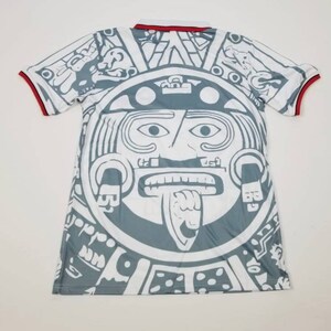 Op de afbeelding: Wit en grijs shirt met korte mouwen met een groot Azteeks kalenderontwerp op de voor- en achterkant. Het shirt heeft een witte kraag en rode biesjes op de mouwen. Het ontwerp is een gedetailleerde illustratie van een Azteekse zonnesteen.