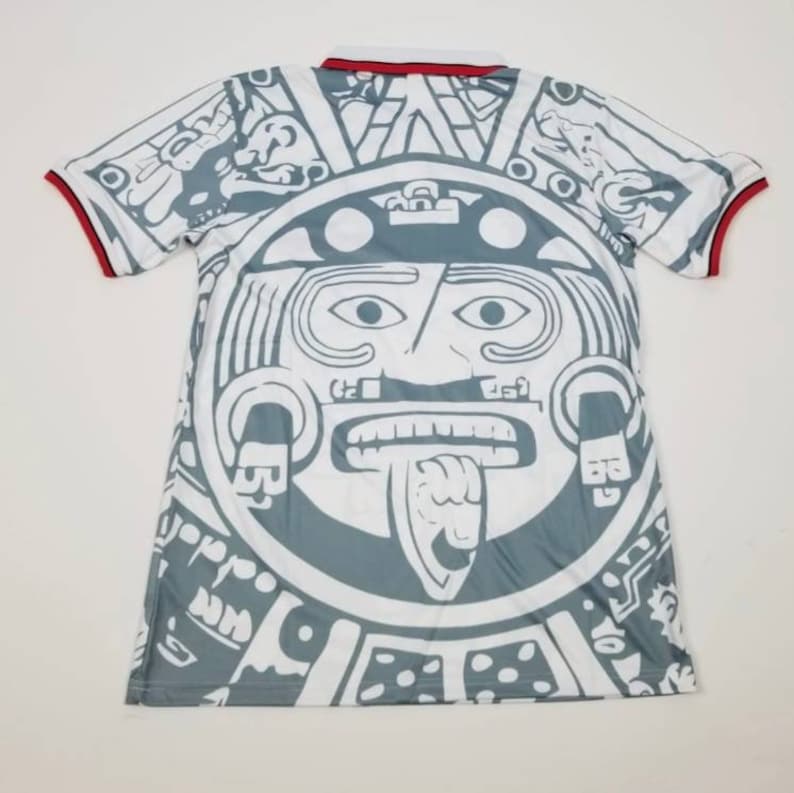 Op de afbeelding: Wit en grijs shirt met korte mouwen met een groot Azteeks kalenderontwerp op de voor- en achterkant. Het shirt heeft een witte kraag en rode biesjes op de mouwen. Het ontwerp is een gedetailleerde illustratie van een Azteekse zonnesteen.