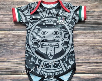 May include: Black and white baby onesie with a gray Aztec-inspired design. The onesie has red and green trim on the sleeves and the text "MEXICO" and "COPA MUNDIAL FIFA FRANCIA '98" on the front.
