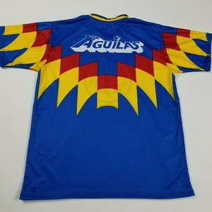 May include: A blue soccer jersey with yellow, red, and white accents. The back of the jersey has the word "AGUILAS" in white with a star above it.