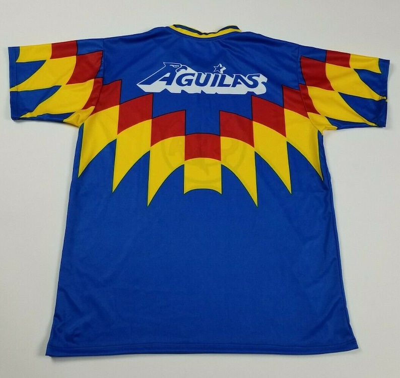 May include: A blue soccer jersey with yellow, red, and white accents. The back of the jersey has the word "AGUILAS" in white with a star above it.